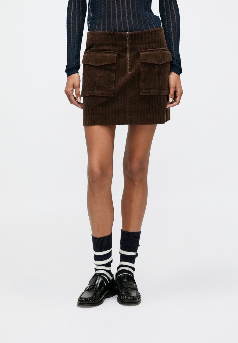 Jupe en velours côtelé marron avec fermeture éclair sur le devant, deux grandes poches et une taille ajustée. Associée à des chaussettes rayées bleu marine et blanches et des mocassins noirs.