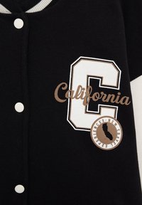 Veste varsity noire avec boutons blancs, col rayé et texte beige « California » sur un grand « C » blanc avec un patch rond montrant l'État de Californie et « New Travel Experiences ».