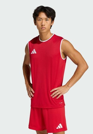 Giovane uomo in piedi con le mani sui fianchi, indossa una maglietta sportiva rossa senza maniche e pantaloncini rossi abbinati con loghi bianchi su sfondo semplice.