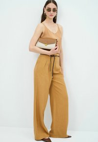 Tan mouwloos topje, mosterdgele wijde broek met plooien, bruine en crèmekleurige handtas met gouden sluiting, en bruine puntige flats.