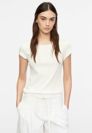 Młoda kobieta o długich brązowych włosach, ubrana w biały krótki rękaw i białe spodnie z wysokim stanem, ręce w kieszeniach na jednolitym tle.