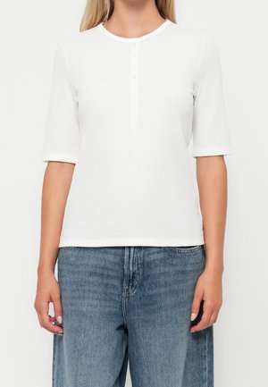 Vrouw die een witte geribbelde korte mouwen henley-shirt en een high-waisted blauwe spijkerbroek draagt, staande tegen een effen achtergrond.