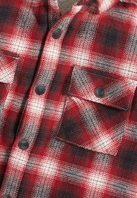 Czerwona, czarna i biała koszula flanelowa w kratę z zatrzaskami i zapinaną na guzik kieszonką na piersi, ukazująca teksturę materiału i detale szwów.