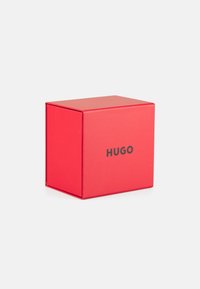 Caja de cartón rojo con acabado mate, con la palabra "HUGO" en una fuente negra en negrita en la parte frontal. La caja tiene bordes afilados y una tapa de levantamiento.