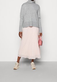 Persoon in een grijze coltrui, lichte roze plooien midi-rok, beige sneakers, en met een kleine roze handtas.