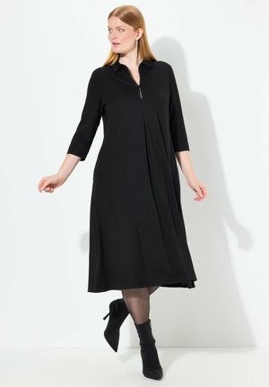 Robe noire zippée avec col, manches trois quarts et design plissé, associée à des bottines noires et des collants transparents.