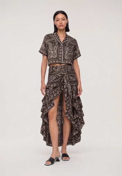 Femme portant un crop top marron à manches courtes imprimé paisley et une jupe à volants asymétrique avec une fente à l’avant, associée à des sandales à talons noires.