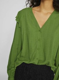 Green, textured blouse with a v-neck and button front. Features ruffled accents on sleeves. Paired with a black, floral lace skirt.