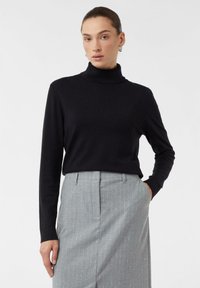 Frau mit zurückgegelten Haaren, die silberne Ohrhänger trägt, einen schwarzen Rollkragenpullover und einen hellgrauen Nadelstreifenrock hat, mit der Hand in der Tasche.