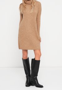 Abito in maglia color marrone chiaro con colletto alto e maniche lunghe, abbinato a stivali neri in pelle al ginocchio con zip laterale.