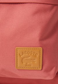 Mochila de tela texturizada rosa con un parche de cuero marrón claro que presenta un cocodrilo y el texto "LACOSTE PARIS". Detalle de costura suave.