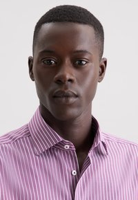 Junger schwarzer Mann mit kurzen Haaren, der ein lila-weiß gestreiftes Hemd mit Knöpfen trägt und direkt in die Kamera blickt, vor einem schlichten Hintergrund.
