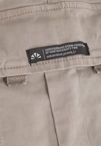 Beige denimjeans med en strukturerad yta. Har en svart etikett med vit text, sydda detaljer och en knappstängning.