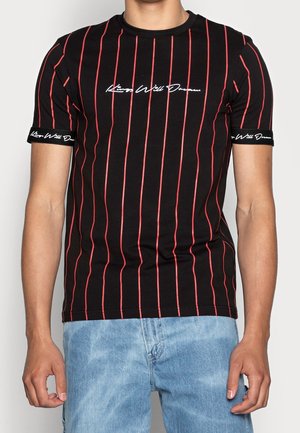 Homme portant un t-shirt noir à manches courtes avec des rayures verticales rouges, du texte en écriture blanche sur la poitrine et les poignets, assorti à un jean en denim bleu clair.