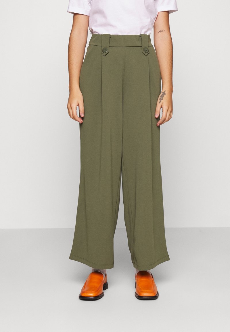 Pantalon à jambes larges en tissu vert olive, avec une taille haute, des accents à boutons et une texture lisse, associé à des chaussures orange.
