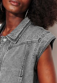 Veste en jean grise sans manches, dotée de clous argentés sur les épaules, d'un col classique, d'un tissu texturé et de boutons sur le devant.