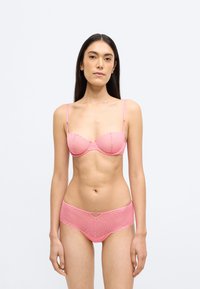 Rožinės spalvos nėriniuota liemenėlė su banguotais kraštais ir reguliuojamomis petnešėlėmis, derančiomis su to paties rašto geometriniu raštu ir mažais dekoratyviniais elementais pasižyminčiomis trumpomis kelnaitėmis.