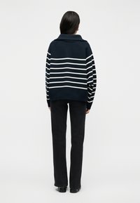 Femme aux longs cheveux noirs portant un pull rayé bleu marine et blanc, un pantalon noir et des chaussures noires, debout de dos sur un fond blanc.