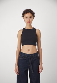 Juodas trumpas tankas su raukšlėtu tekstūru, apvaliu iškirpimu ir be rankovių dizainu, derinamas su aukštu liemeniu tamsiais džinsais.