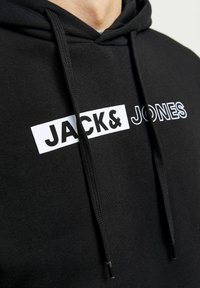 Čierna mikina s kapucňou s bielou grafikou a textom "JACK&JONES." Vyrobená z mäkkého materiálu, s nastaviteľnou šnúrkou a rebrovanými manžetami.
