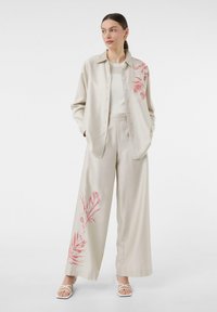 Jasno beżowa koszula z guzikami z czerwonym haftem kwiatowym; zestawiona z szerokimi beżowymi spodniami, tworząc skoordynowaną stylizację. Białe sandały.