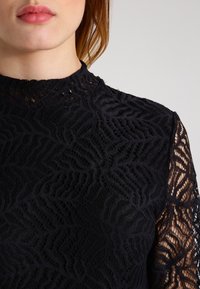 Svart spetsblus med hög halsringning och långa ärmar med blommönster, kontrasterande genomskinliga detaljer på ärmarna.