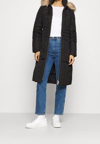 Svart quiltad pufferjacka med dragkedja framtill och huva med pälsdetalj, bärs över en vit skjorta och blå denimjeans. Vita sneakers fulländar looken.