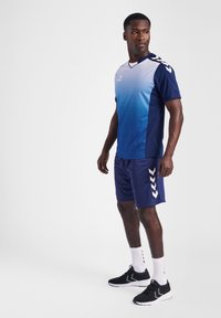 Blauw-wit sportshirt met een V-hals, met een verloopontwerp en geometrisch patroon; gecombineerd met blauwe shorts en zwarte sportschoenen.