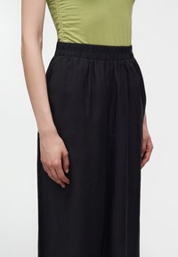 Femme portant un haut vert froncé rentré dans un pantalon noir ample taille haute avec une ceinture élastique, mains détendues sur les côtés.