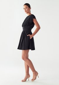 Black dress with short sleeves, fitted waist, and gathered skirt. Features pockets and paired with beige strappy high heels.