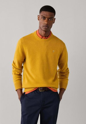 Ein Mann steht mit den Händen in den Taschen, trägt einen gelben Pullover über einem orangefarbenen Hemd und eine marineblaue Hose, blickt geradeaus vor einem schlichten Hintergrund.