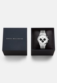 Orologio Daniel Wellington argento con quadrante bianco e sottoquadranti neri, in scatola nera accanto al coperchio blu navy con nome del brand in oro.