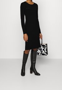 Robe noire à manches longues avec une silhouette ajustée et des découpes courbes, associée à des bottes en cuir noires montant jusqu'au genou et un sac à main noir et blanc à motifs.