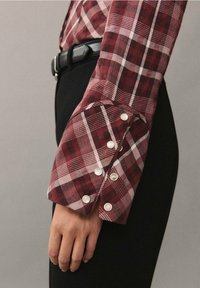 Červená a bílá kostkovaná košile s širokým manžetovým lemem a třemi bílými knoflíky, v kombinaci s černými kalhotami. Zaměření na texturu tkaniny a vzory.