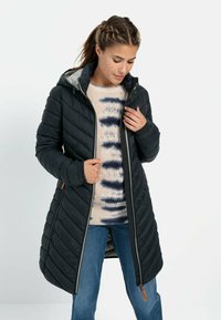 Zwarte gewatteerde lange jas met een capuchon, voorzien van een ritssluiting, grijze voering en gestructureerde stof, gedragen boven een beige trui met blauwe patronen.
