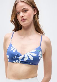 Νεαρή γυναίκα που φοράει ένα μπλε μπραλέτο με λουλουδάτο σχέδιο και λεπτές τιράντες, πάνω σε απλό ανοιχτό φόντο.