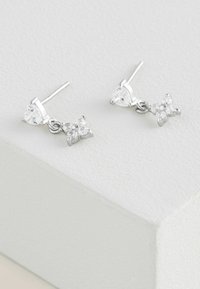 Orecchini a stud in argento con una parte superiore a forma di cuore e un dettaglio pendente a forma di fiocco, impreziositi da pietre chiare e scintillanti.