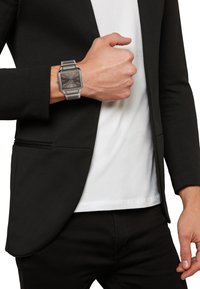 Sølv rektangulær ur på et metal link armbånd, med en mørk skive og sølvmarkeringer, båret på et håndled mod en sort blazer og hvid skjorte.