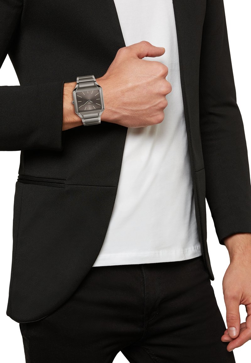 Sølv rektangulær ur på et metal link armbånd, med en mørk skive og sølvmarkeringer, båret på et håndled mod en sort blazer og hvid skjorte.