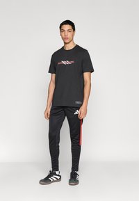 Μαύρο βαμβακερό t-shirt με το γράφημα "BORN TO SCORE", συνδυασμένο με μαύρο παντελόνι φόρμας με κόκκινες ρίγες και λογότυπο με τρεις ρίγες. Μαύρα αθλητικά παπούτσια.