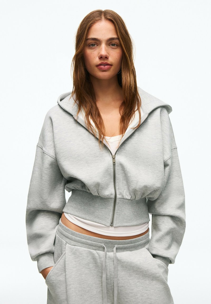 Jeune femme aux cheveux bruns portant un sweat à capuche court gris clair zippé et un pantalon de survêtement assorti, les mains dans les poches.