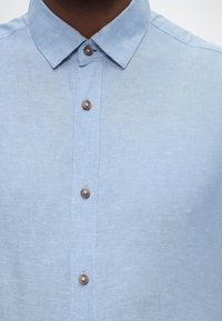 Ανοιχτό μπλε πουκάμισο με σκούρα καστανά κουμπιά και κανονικό γιακά, φορεμένο από άτομο που απεικονίζεται από τον λαιμό μέχρι το μέσο του στήθους.