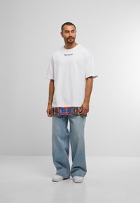 Mann steht da, trägt ein übergroßes, weißes T-Shirt mit buntem Saum, helle, weite Jeans und weiße Sneakers vor einem schlichten grauen Hintergrund.