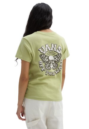Person mit langen dunklen Haaren trägt ein hellgrünes Vans T-Shirt mit Totenkopf- und Schmetterlingsmotiv und weiße Hosen, von hinten gezeigt.