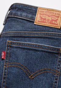 Tmavomodré džínsy z denimu s koženou nášivkou na páse, s oranžovým prešívaním a malou červenou nálepkou s nápisom "Levi's" na zadnej vrecku.