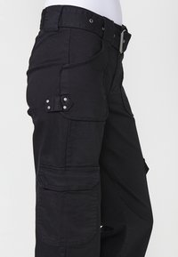 Pantalones cargo negros con un diseño ajustado, que presentan un cierre de botón, múltiples bolsillos y acentos de herrajes en plata. Textura suave.