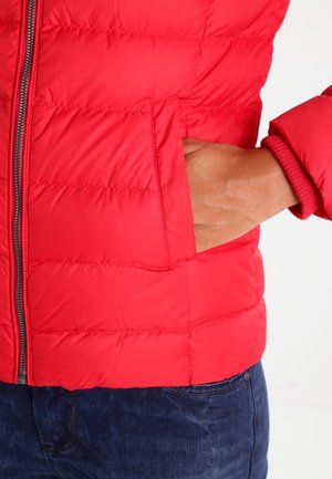 Main dans la poche d'une veste matelassée rouge avec une fermeture éclair visible et un jean bleu en dessous.
