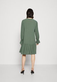 Vestido verde con mangas largas, detalles plisados y una falda acampanada. Tejido ligero, de media longitud. Combinado con zapatos de tacón alto negros.