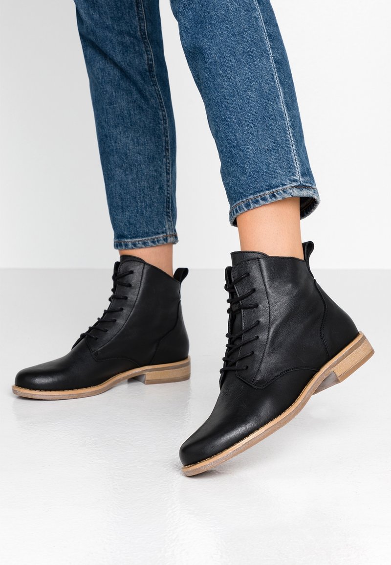Personne portant des bottines en cuir noir à lacets, associées à un jean bleu retroussé, debout sur un sol clair devant un fond blanc.