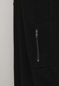 Čierny textilný odev s vreckom na zips. Textúra sa zdá byť mierne pokrčená, so hladkým zipsom na vrecku.